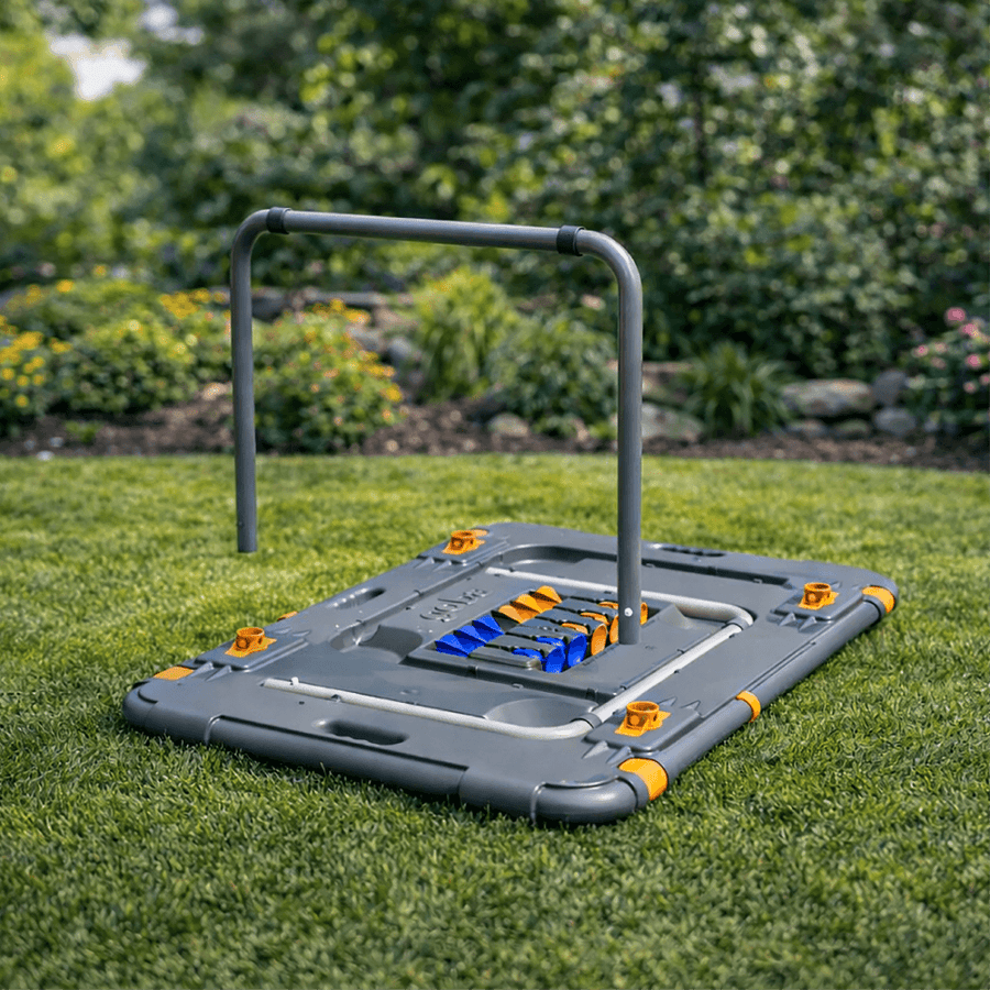 A gray plastic fitness trampoline pad with a black metal handlebar frame and orange/yellow corner bumpers, shown on a green grass lawn with a blurred garden background.